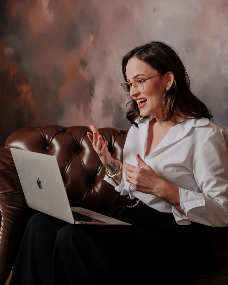 Fernanda Fontanetti conversando e gesticulando, com o notebook no colo, em uma poltrona de couro